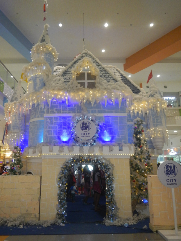 Ein festliches Weihnachtsdorf mit einer burgenähnlichen Struktur, die mit Lichtern geschmükt ist, umgeben von Menschen in einem Einkaufszentrum mit beleuchteter Decke und einem Textboard auf der rechten Seite.