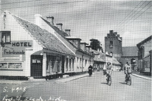Ein Schwarz-Weiß-Foto von Menschen, die auf Fahrrädern eine Straße entlangfahren, die von Gebäuden, Bäumen und Drähten gesäumt ist, mit Text am unteren Rand des Bildes.