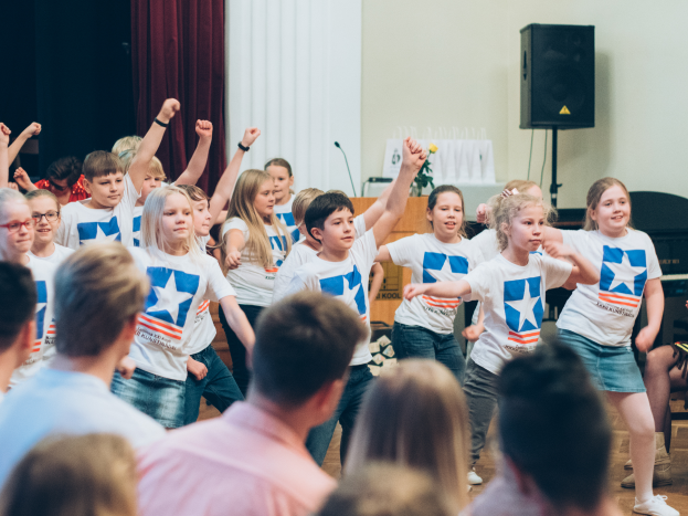 Eine Gruppe von Kindern in weißen Hemden und blauen Shorts tanzt vor einem sitzenden Publikum, mit einer Vorhangwand, Lautsprecherständer und einem Tisch mit Mikrofon im Hintergrund.