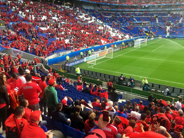 Eine große Menschenmenge in einem hell erleuchteten Stadion schaut einem Fußballspiel zu, mit Zuschauern auf Sitzplätzen und Stehplätzen, Fahnen, einem Torpfosten und Deckenleuchten.