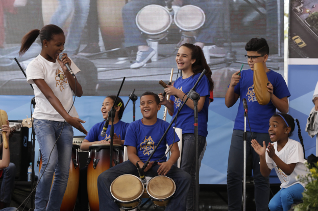 Kinder, die auf einer Bühne Musikinstrumente spielen, mit einer Frau in der Mitte, die ein Mikrofon hält, umgeben von Mikrofonstativen, einem Banner im Hintergrund und Pflanzen auf der rechten Seite.