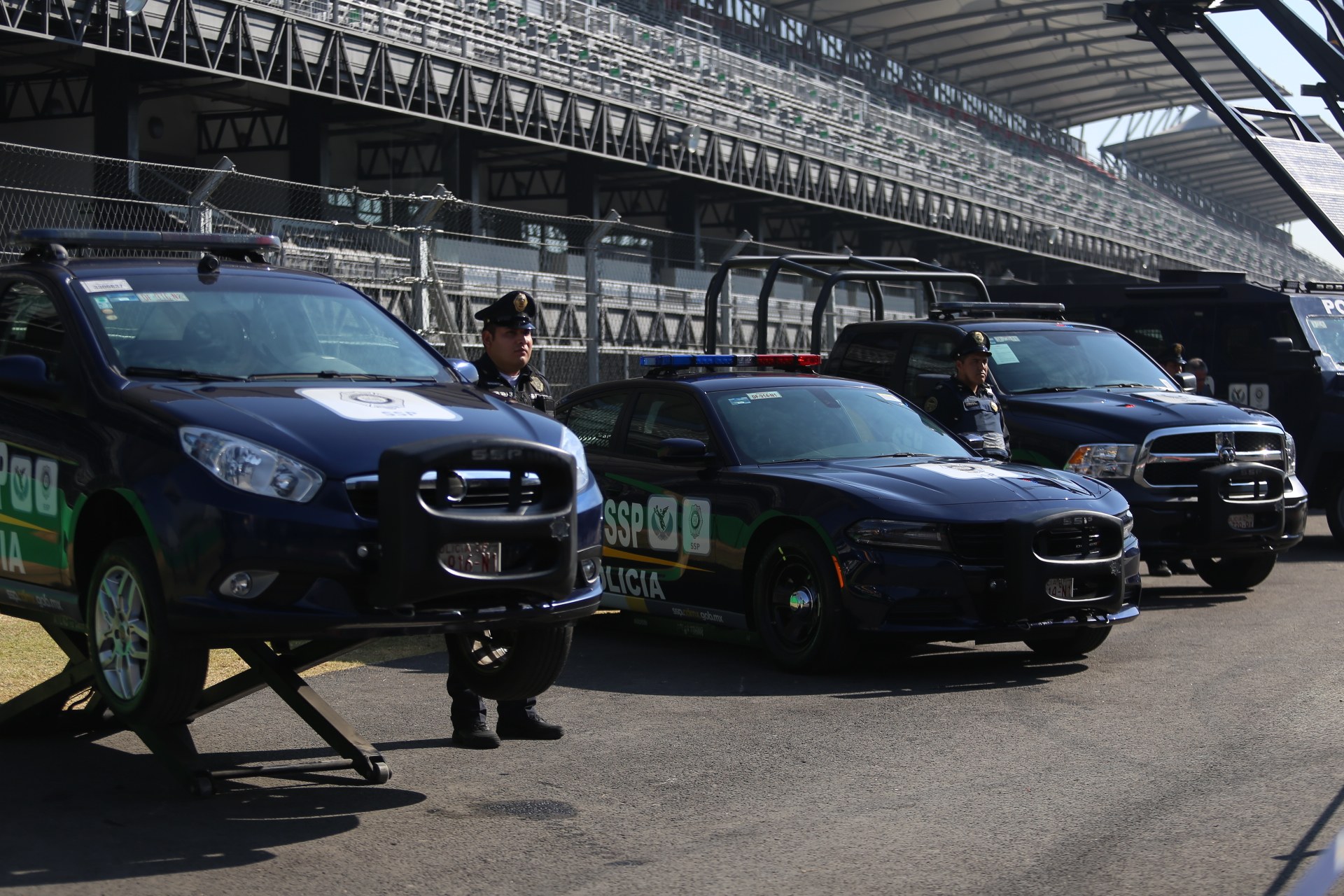 Eine Gruppe von Polizeiwagen steht vor einer Rennstrecke, uniformierte Beamte stehen auf der Straße, im Hintergrund ein Stadion und Maschendrahtzaun.
