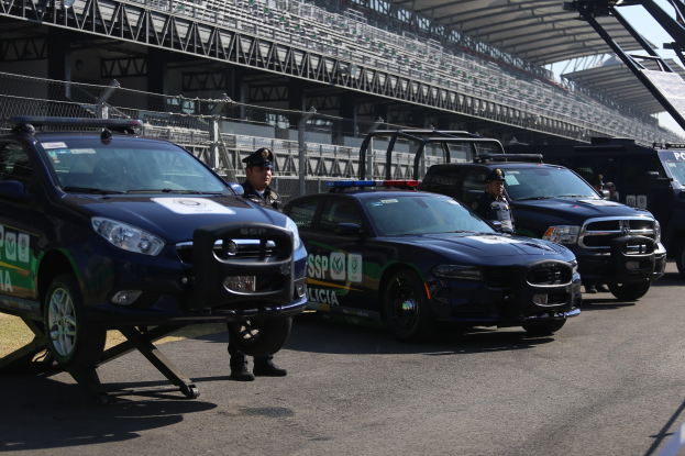 Eine Gruppe von Polizeiwagen steht vor einer Rennstrecke, uniformierte Beamte stehen auf der Straße, im Hintergrund ein Stadion und Maschendrahtzaun.