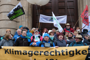 Eine Gruppe von Menschen steht vor einem Gebäude mit Säulen und einer Tür, hält ein Transparent mit Text, Fahnen mit Stangen und Ballons und nimmt an einem Klimastreik in Deutschland teil.