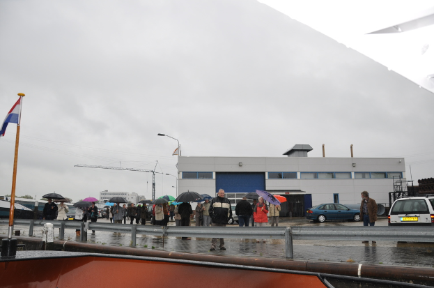 Eine Gruppe von Menschen, die draußen stehen, einige halten Schirme, mit einer Fahne an einem Mast, zwei Autos, einem Gebäude mit Fenstern, einem Turmkran, einer Straßenlaterne und einer Absperrung.