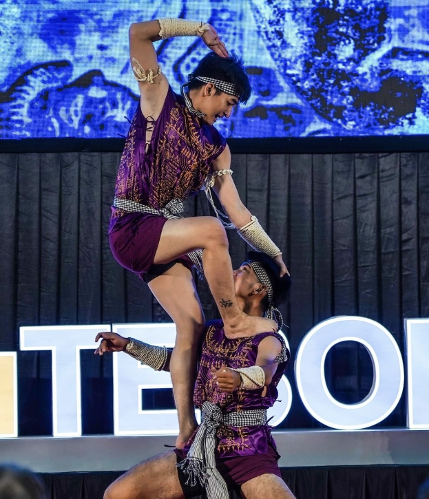 Zwei Akrobaten führen auf der Bühne eine Darbietung auf, wobei der Mann die Frau in seinen Armen hält, während beide purpurne Kleider tragen, und ein Publikum sowie ein Bildschirm im Hintergrund zu sehen sind.