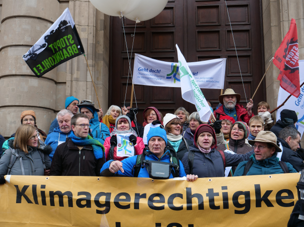 Eine Gruppe von Menschen steht vor einem Gebäude mit Säulen und einer Tür, hält ein Transparent mit Text, Fahnen mit Stäben und Ballons und nimmt an einem Klimastreik in Deutschland teil.