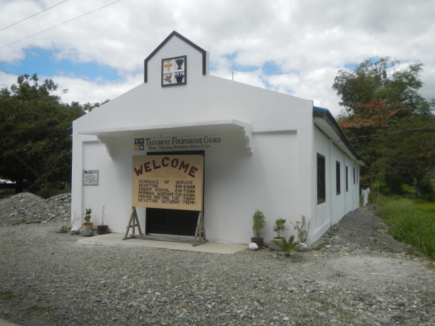 Kleine weiße Kirche mit einem "Willkommen"-Schild, umgeben von Topfpflanzen, Gras, Bäumen, Stromleitungen und einem bewölkten Himmel.