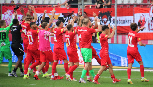 Gruppe von Fußballspielern, die ein Tor feiern, mit Flaschen oder klatschenden Händen, Bannern, einem Zaun, Metallstangen, Stühlen und einem Dach mit Lampen im Hintergrund.