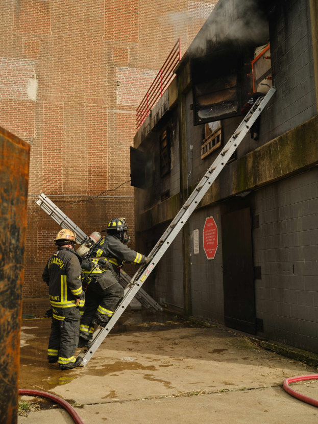 Feuerwehrleute mit Helmen, die eine Leiter zu einem raucherfüllten Gebäude hinaufklettern, mit Metallobjekten auf der linken Seite und Rohren auf dem Boden.