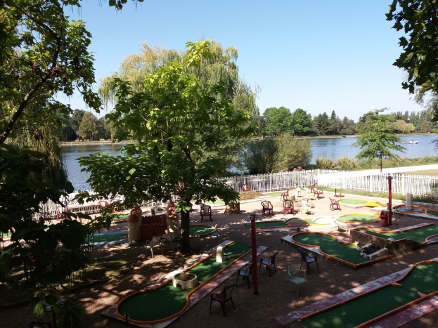 Minigolfanlage in einem Park mit grünem Gras, Bäumen und einem Zaun, umgeben von verstreuten Stühlen und Tischen, mit einem Gewässer und einem Boot im Hintergrund unter einem sichtbaren Himmel.