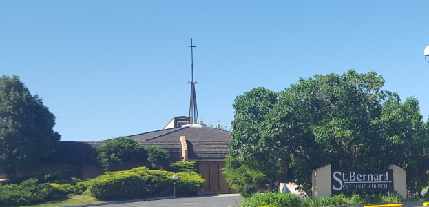 St. Bernard Catholic Church in San Antonio, Texas, ein Gebäude mit einem Kreuz darauf, umgeben von Pflanzen, Gras, Bäumen, einem Straßenpfahl, einer Schautafel, Straßenlaternen und einem bewölktem Himmel.