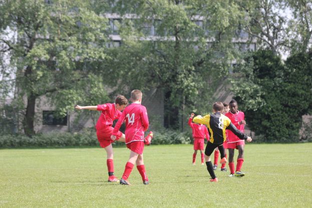 Fünf Jungs in roten T-Shirts und Shorts spielen Fußball auf einem grasbewachsenen Feld mit Bäumen und Gebäuden im Hintergrund.