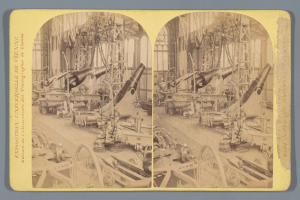 Ein altes Schwarz-Weiß-Foto einer Fabrik mit einem großen Geschütz in der Mitte, umgeben von mehreren anderen Geschützen, Metallstangen und verschiedenen Gegenständen auf dem Boden, mit Text auf der linken Seite.