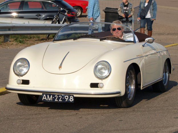Ein Mann fährt einen weißen Porsche 356 Speedster über eine Straße, umgeben von Menschen in verschiedenen farbigen Kleidern, mit einem Geländer und einem schwarzen Müllimer an der Seite und weiteren Autos und einem Gebäude im Hintergrund.