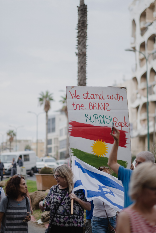 Eine Gruppe von Menschen marschiert nachts die Straße entlang und hält ein Schild hoch, auf dem steht: "Wir stehen zu den tapferen kurdischen Menschen", mit einem geparkten Van, Straßenlaternen, Bäumen und Gebäuden im Hintergrund.