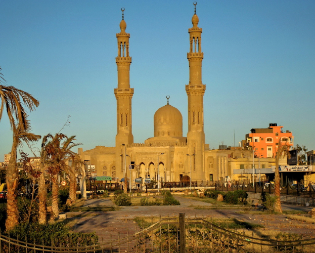 Eine Moschee ist zentral mit Gebäuden im Hintergrund, Bäumen auf beiden Seiten, einem Tor, einem Weg, Pflanzen, Pfählen, Brettern, Fahrzeugen, einem Gebäude mit einem Geländer auf der rechten Seite und Himmel oben.