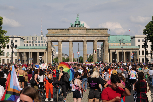 Eine große Menschenmenge marschiert vor dem Brandenburger Tor in Berlin, hält Fahnen und Banner hoch und einige fahren Fahrräder; die Straße ist mit Laternenmasten und Bäumen gesäumt und der Himmel ist bewölkt, im Hintergrund sind Gebäude mit Fenstern und Bögen sowie eine Statue zu sehen.
