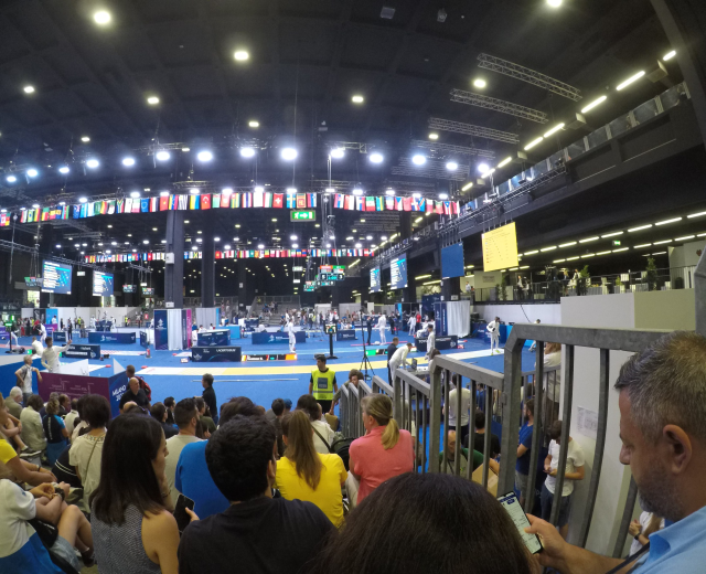 Eine große Menschenmenge sitzt und steht in einer Indoor-Sportarena, mit einem Metallzaun auf der rechten Seite, Bildschirmen, Flaggen und Deckenlampen im Hintergrund.
