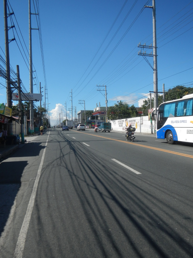 Ein Bus fährt auf einer Straße mit hohen Strommasten und Kabeln, flankiert von Geschäften und Fußgängern auf der linken Seite, mit einer Wand, Bäumen, Gebäuden und einem bewölkten Himmel im Hintergrund.