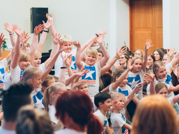 Eine Gruppe von Kindern steht mit erhobenen Händen vor einer Menge, mit einem Tisch, der eine Vase und einen Lautsprecher hält, links und einer Wand mit einer Tür im Hintergrund, was eine Sommercamp-Atmosphäre in Schweden vermuten lässt.