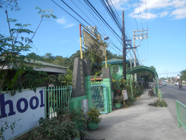 Einfahrt zu einer Schule mit einem "Schule"-Schild, Fahrzeuge auf der Straße, ein Tor, ein Tor, eine Namensschild, Pflanzen, Bäume, Strommasten mit Drähten und einen bewölkten Himmel im Hintergrund.