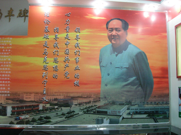 Großes Mao-Zedong-Plakat an einer Gebäudewand, beleuchtet von Lampen, mit einem Foto-Rahmen und Text rechts daneben.