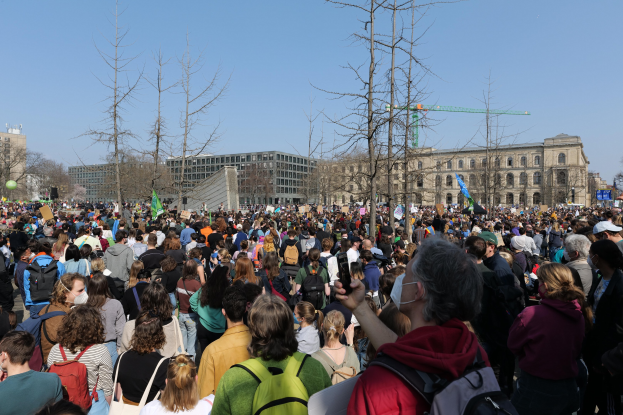 Eine große Menge Menschen mit Schildern und Taschen vor einem Gebäude mit Fenstern, Bäumen und einem klaren Himmel im Hintergrund, was auf eine Klimaprotestdemo hinweist.