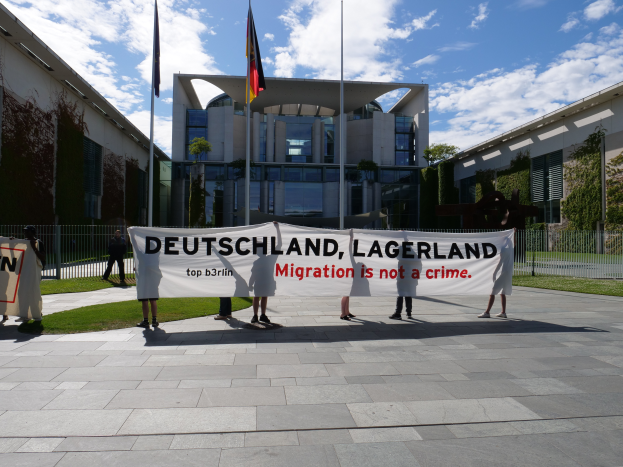 Eine Gruppe von Menschen hält ein Transparent mit der Aufschrift "Deutschland, Lagerland Migration ist kein Verbrechen" vor einem Gebäude mit Fenstern, umgeben von Gras, einem Metallzaun, Pflanzen, Bäumen und Fahnen auf Ständen, unter einem bewölkten Himmel.
