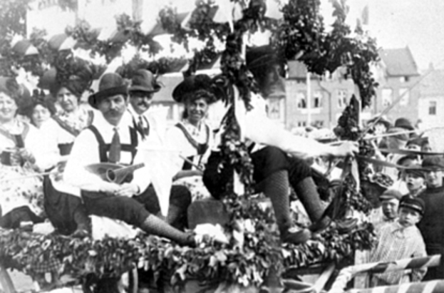 Eine Gruppe von Menschen in traditioneller bayrischer Kleidung fährt auf einem mit Laub geschmückten Festwagen in einem Umzug, einige halten Musikinstrumente, mit Gebäuden und einem klaren Himmel im Hintergrund, dargestellt in Schwarz-Weiß.