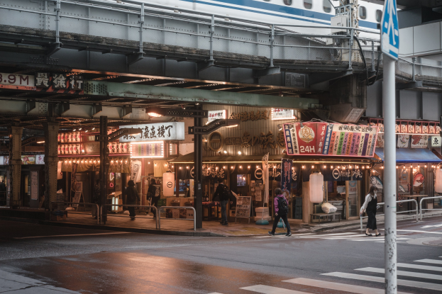 Menschen überqueren eine Straße neben einem Zug, mit einem Hinweisschild im Vordergrund und Gebäuden, Schildern und einer Brücke mit einem Zug im Hintergrund.