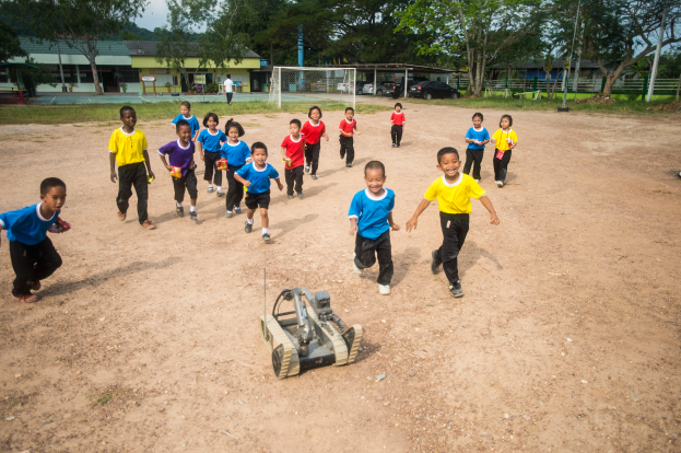 Eine Gruppe von Kindern, die auf einem Schotterfeld Fußball spielen, umgeben von Bäumen, Gebäuden, Pfählen, Fahrzeugen, Netzen und anderen Gegenständen, mit einem Fahrzeug im Vordergrund und dem Himmel im Hintergrund.