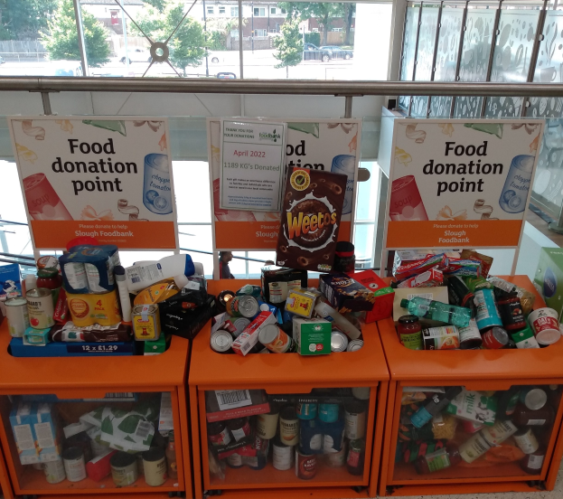 Orange Schübe mit Lebensmittel wie Flaschen, Dosen und Kartons dienen als Spendenstelle, mit Texttafeln und einem Blick auf Fahrzeuge, Bäume, Gebäude und Himmel durch Glasfenster.