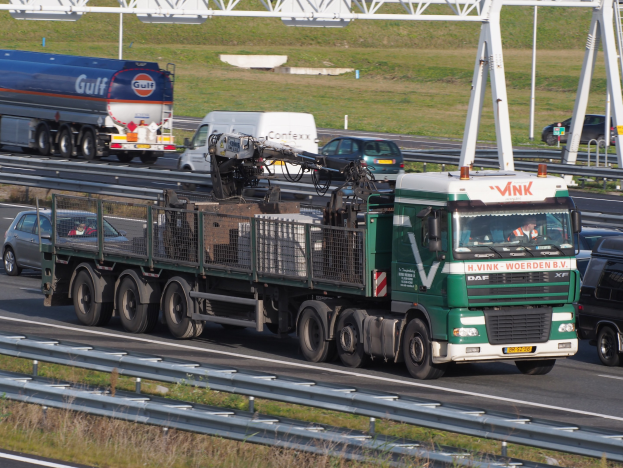 Ein großer Lastwagen mit einem Anhänger fährt auf einer Straße neben einer Brücke, mit Gras und Geländern auf beiden Seiten, unter einem klaren blauen Himmel mit Masten im Hintergrund.