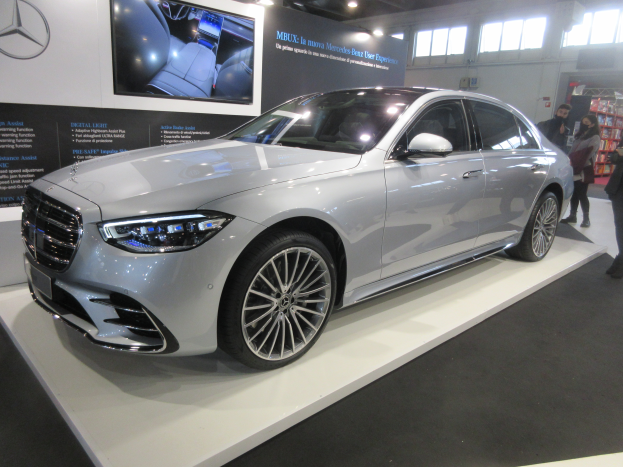 Ein Mercedes-Benz S-Klasse-Saloon wird auf der Frankfurter Motor Show präsentiert, umgeben von Menschen, mit einer Texttafel und einem Logo auf der linken Seite und Lichtern, Fenstern und anderen Objekten im Hintergrund.