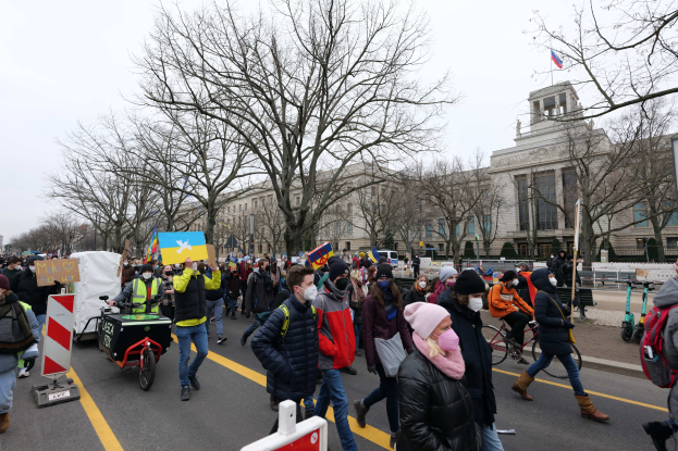 Eine große Protestmarsch mit Menschen, die Schilder halten und Fahrräder fahren, auf einer Straße in Washington, D.C., mit Bäumen, Schildern und einem klaren blauen Himmel im Hintergrund.