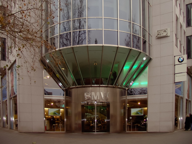 Großes BMW-Hauptquartier-Gebäude in Frankfurt, Deutschland, mit Glaswänden, Text an der Fassade, Menschen vor dem Gebäude und einem Baum links.