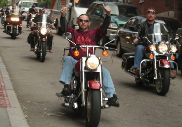 Eine Gruppe von Menschen, die auf Fahrrädern auf einer Straße fahren, wobei die Person in der Mitte ein rotes Hemd trägt und ohne Hände fährt, und eine andere Person rechts mit einer schwarzen Jacke und einem Lächeln. Im Hintergrund sind zwei Autos und ein Van zu sehen.