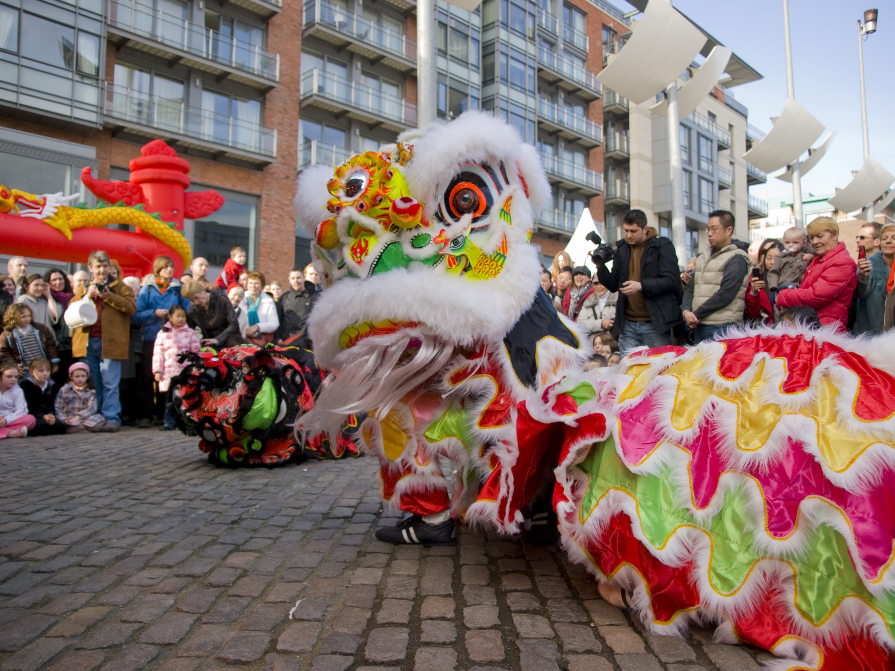 Ein lebendiges chinesisches Neujahrsfest in Amsterdam mit einem Löwen tanzen im Vordergrund und einer Menschenmenge, einige halten Kameras, während im Hintergrund Gebäude und Laternenpfähle zu sehen sind, unter einem klaren blauen Himmel.