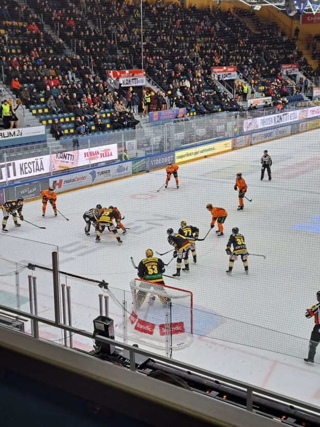 Eine Gruppe von Menschen, die Hockey auf einem Eisstadion spielen, umgeben von einem Zaun, mit Zuschauern im Hintergrund und beleuchteten Stadionlampen oben.