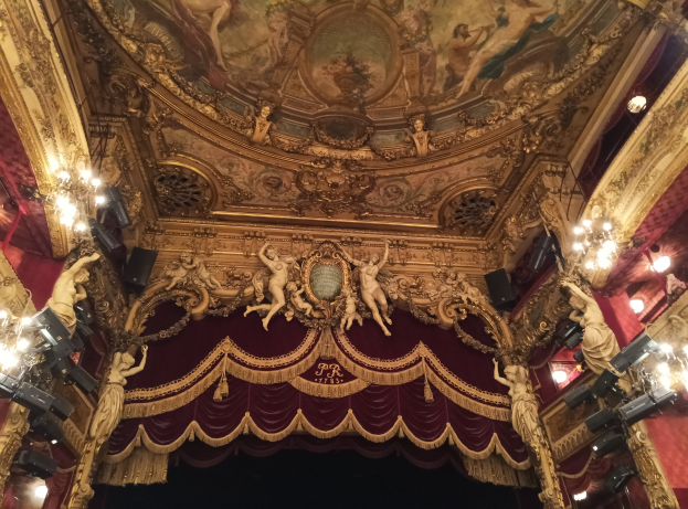 Theaterinnenraum mit roter Gardine, Kronleuchtern, Deckenplastiken, Lichtern und einem Gemälde, das eine Bühne mit einigen Menschen am Boden zeigt.