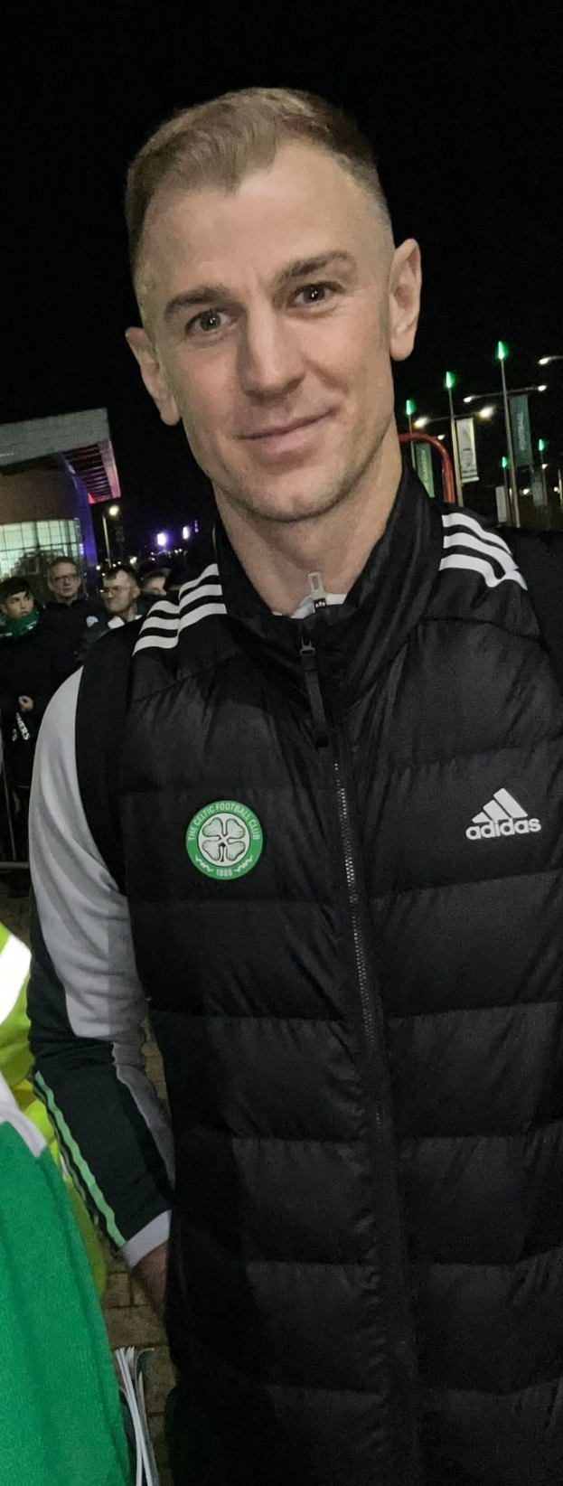 Ein Mann in einer schwarzen Jacke steht lachend vor einer Menge, beleuchtet von Gebäudelichtern und dem Himmel, wahrscheinlich feiernd nach Celtic's Sieg im schottischen Cup-Finale.