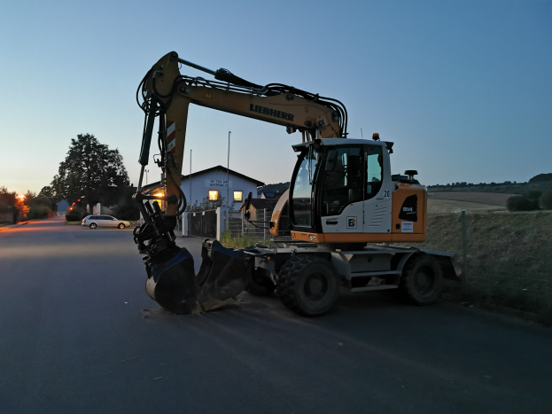 Ein Liebherr LTM 1100-5 Bagger neben einer Stra├če geparkt, umgeben von Gras, einem Zaun, B├Ąumen, H├Ąusern, Masten und einem bew├Âlktem Himmel.