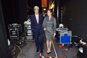 Angelina Jolie und John Kerry auf einem Boden umgeben von einer Gruppe von Menschen, verschiedenen Geräten, Kabeln, einem Helm auf einem Wagen, einem Müllcontainer, einem Vorhang, einer Säule und einer Decke mit Lampen.