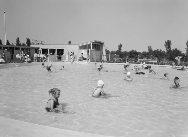 Schwarzes und weißes Foto von Kindern, die in einem Swimmingpool mit Gebäuden, Bäumen, Pfählen, Pflanzen und einem Zaun im Hintergrund spielen.