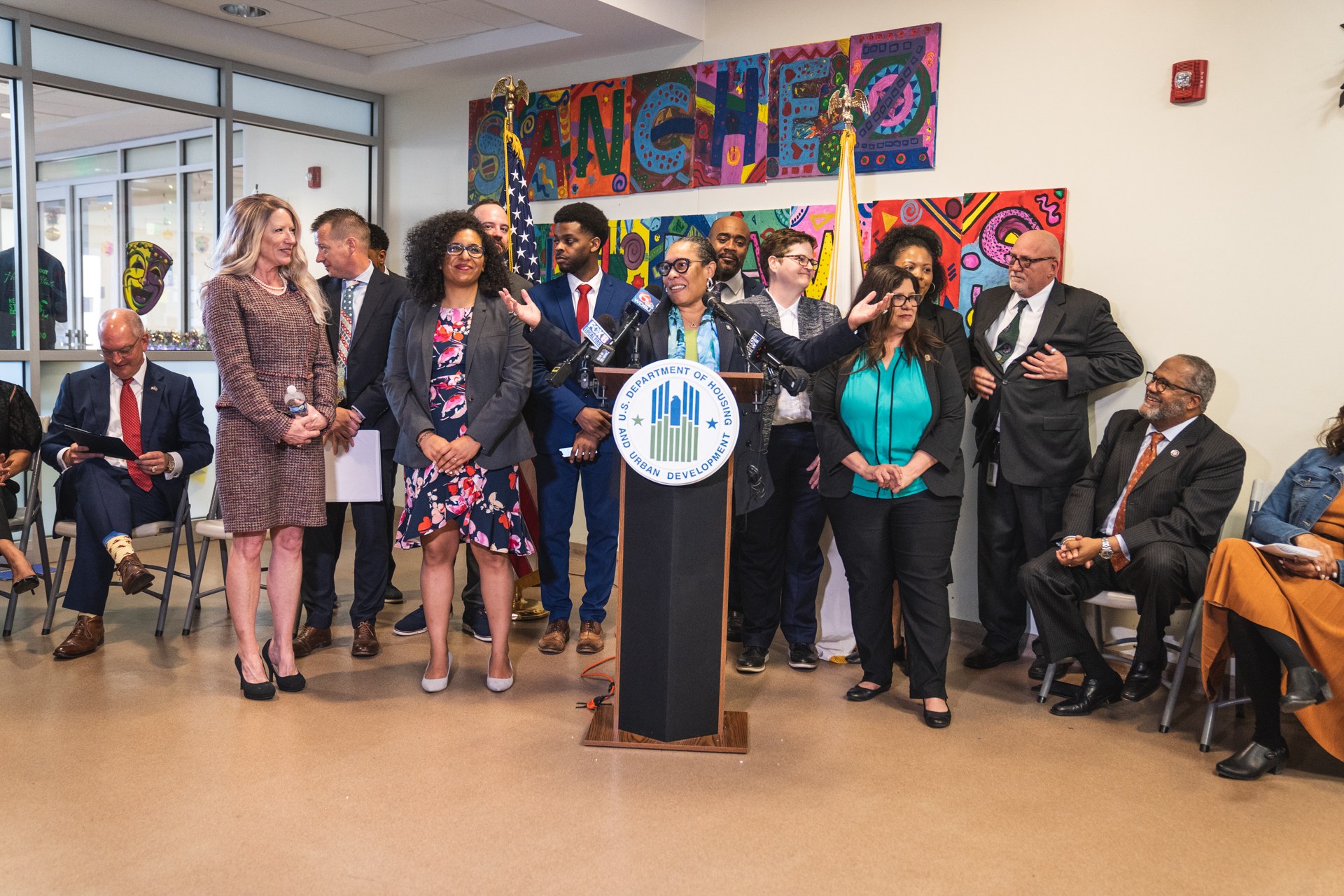 Eine Gruppe von Menschen steht in der Nähe eines Podiums mit einem Mikrofon und einer Tafel, auf der "Sancho Stadtverordnetenrat tritt dem Stadtrat bei" steht, während andere auf Stühlen sitzen oder in der Nähe stehen; Fahnen und ein Feuerlöscher sind an der Rückwand zu sehen.