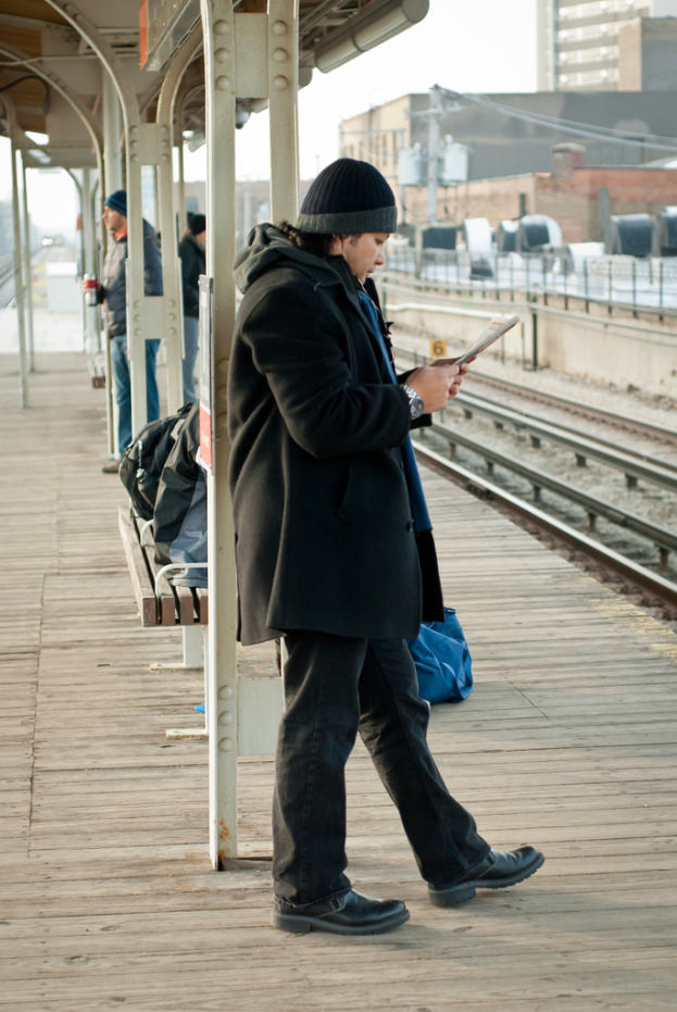 Ein Mann in schwarzer Jacke steht an einem Bahnhof mit einer Eisenstange und einem Schuppen hinter ihm und Eisenbahnschienen und braune Gebäude rechts von ihm.
