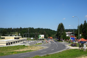 Ein Kreisverkehr mit Fahrzeugen, Gras, Laternenmasten, Texttafeln, Gebäuden, Bäumen und einem bewölkten Himmel, mit einem Straßenschild im Fokus.
