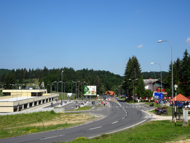 Ein Kreisverkehr mit Fahrzeugen, Gras, Laternenmasten, Texttafeln, Gebäuden, Bäumen und einem bewölkten Himmel, mit einem Straßenschild im Fokus.